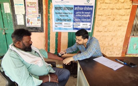 A skin check-up camp at Umrale: a dermatologist examining a patient's arm with a hand lens under the Project Arogyam banner.