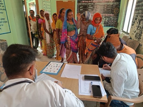 A village OPD in a panchayat hall — women waiting in line while the doctor consults and a colleague enters each case into the record.